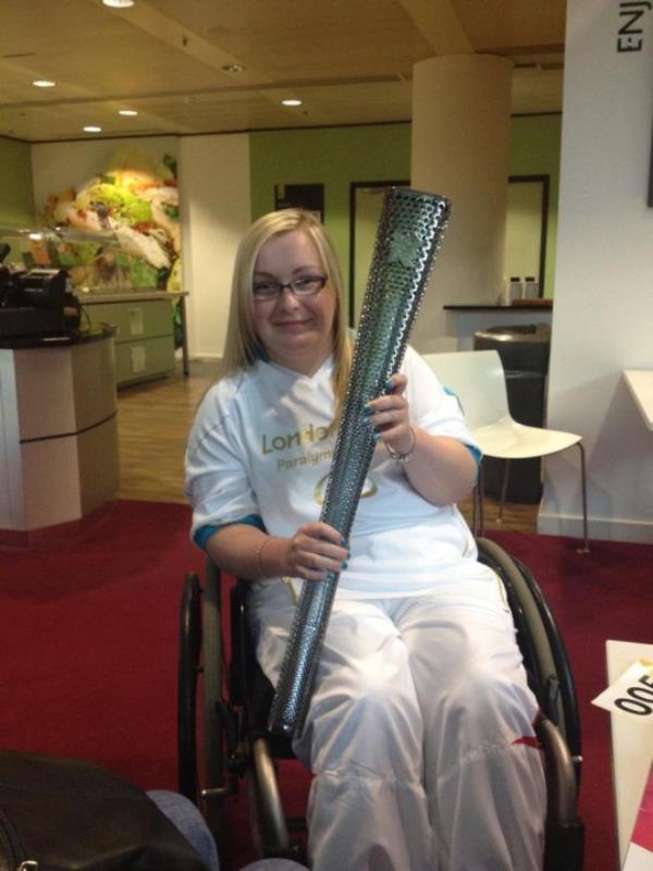 Maisie holding the Olympic torch, in her wheelchair, in an office with a red carpet. She is wearing all white, has blo<em></em>nde hair and black glasses.